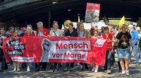 Straßenszene unter einer Brücke mit einer großen Gruppe gehender Personen, die ein breites rotes Banner tragen. Auf dem Banner steht: „1. Mai 2026 in Köln“, „Jetzt erst recht!“, „Mensch vor Marge“ und „DGB“. Weitere Schilder und Fahnen sind im Hintergrund zu sehen, darunter ein Schild mit der Aufschrift „Zukunft sichern statt Sozialstaat schwächen. Stoppt den KAHLSCHLAG“.