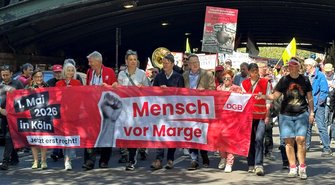 Straßenszene unter einer Brücke mit einer großen Gruppe gehender Personen, die ein breites rotes Banner tragen. Auf dem Banner steht: „1. Mai 2026 in Köln“, „Jetzt erst recht!“, „Mensch vor Marge“ und „DGB“. Weitere Schilder und Fahnen sind im Hintergrund zu sehen, darunter ein Schild mit der Aufschrift „Zukunft sichern statt Sozialstaat schwächen. Stoppt den KAHLSCHLAG“.