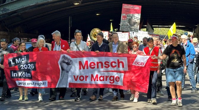 Straßenszene unter einer Brücke mit einer großen Gruppe gehender Personen, die ein breites rotes Banner tragen. Auf dem Banner steht: „1. Mai 2026 in Köln“, „Jetzt erst recht!“, „Mensch vor Marge“ und „DGB“. Weitere Schilder und Fahnen sind im Hintergrund zu sehen, darunter ein Schild mit der Aufschrift „Zukunft sichern statt Sozialstaat schwächen. Stoppt den KAHLSCHLAG“.