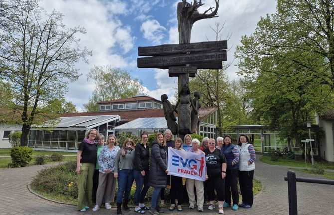 Eine Gruppe von Menschen steht auf einem gepflasterten Weg vor einem hohen, geschnitzten Holzdenkmal mit mehreren quer angebrachten Holztafeln mit eingraviertem Text. Im Vordergrund hält eine Person ein Banner mit der Aufschrift „EVG FRAUEN“. Im Hintergrund sind Bäume, Rasenflächen sowie Gebäude mit Glasfronten und roten Dächern zu sehen.