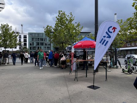 Offener Platz mit mehreren Bäumen, Informationsständen und einer weißen Fahne mit EVG-Logo im Vordergrund. Unter roten und blauen Sonnenschirmen stehen Personen an Tischen. Mehrere Menschen bewegen sich über den Platz. Im Hintergrund stehen moderne Gebäude, Fahrräder und E-Scooter. Der Himmel ist stark bewölkt.