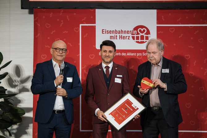 Drei Personen stehen vor einem roten Hintergrund mit dem gut lesbaren Schriftzug „Eisenbahnerin mit Herz“. Die mittlere Person hält eine gerahmte Urkunde, eine andere Person hält ein rotes Objekt in Herzform. Eine Person hält ein Mikrofon. Alle drei tragen Namensschilder.
