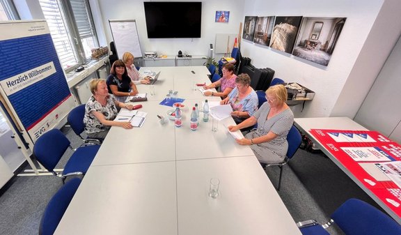 Besprechungsraum mit einem langen weißen Konferenztisch. Sechs Personen sitzen verteilt um den Tisch und arbeiten mit Unterlagen, Notizblöcken und einem Laptop. Auf dem Tisch stehen Wasserflaschen, Gläser und Schreibmaterial. An der linken Seite befinden sich Fenster und ein Flipchart mit dem sichtbaren Text „Herzlich Willkommen“. An der Stirnwand hängen ein großer Bildschirm und mehrere gerahmte Bilder. Rechts liegen rote und blaue Plakate auf einem zusätzlichen Tisch.