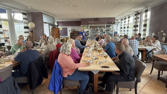 Innenraum eines Restaurants mit mehreren langen Holztischen. Rund drei Dutzend sitzende Personen befinden sich an den Tischen, auf denen Tassen, Gläser, Flaschen und Teller stehen. Im Hintergrund steht eine Person vor dem Thekenbereich. Große Fenster an beiden Seiten lassen Tageslicht in den Raum. An den Fenstern hängen gestreifte Vorhänge in Grau und Weiß. An der Rückwand befinden sich Regale mit Flaschen und Gläsern sowie ein Schild mit der Aufschrift „HOLZER“. Die Decke ist mit vielen kleinen Einbauleuchten ausgestattet.