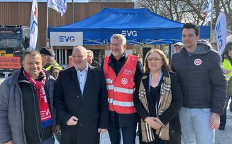 Eine Gruppe von fünf Personen steht im Freien vor einem blauen Pavillon mit EVG‑Logo. Alle blicken Richtung Kamera. Eine Person in der Mitte trägt eine rote EVG‑Warnweste. Hinter der Gruppe sind Fahnen mit EVG‑Aufdruck, ein Lkw sowie Bäume und ein Gebäude zu sehen.