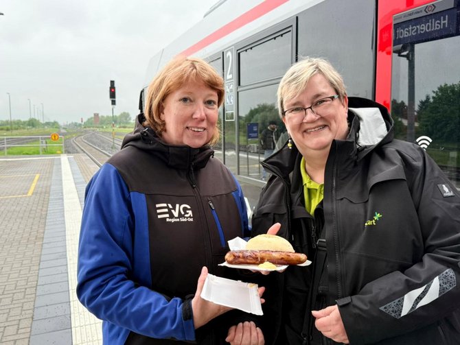 Zwei Personen stehen auf einem Bahnsteig vor einem roten Zug. Eine Person hält eine geöffnete Pappschale mit einem Brötchen und einer Bratwurst. Auf einer Jacke ist der Text „EVG Region Süd-Ost“ zu lesen. Im Hintergrund sind Gleise, Signale und ein Bahnsteig zu sehen.