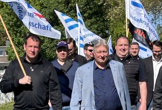 Mehrere Personen stehen im Freien vor grünen Bäumen und halten Fahnen an Holzstangen. Die Fahnen sind überwiegend weiß mit blauen Streifen und tragen gut lesbaren Text wie „Eisenbahn- und Verkehrsgewerkschaft“ sowie „EVG“. Eine weitere Fahne ist schwarz mit orangefarbener Aufschrift „JUGEND“. Die Personen tragen Alltagskleidung wie Jacken, Pullover und Hemden. Im Hintergrund sind weitere Fahnen und Laubbäume zu sehen.