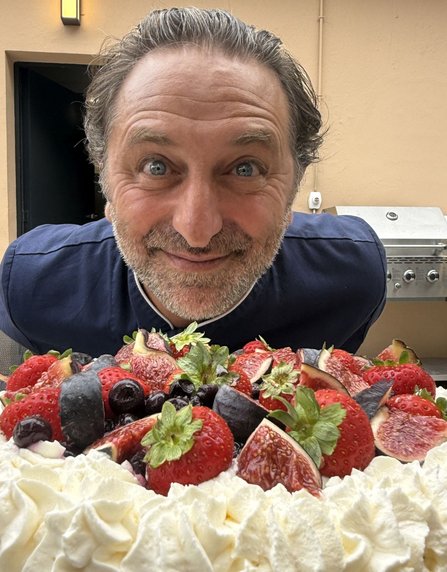 Nahaufnahme einer Person, die sich über einen großen Kuchen beugt. Der Kuchen ist mit weißer Sahne sowie Erdbeeren, Feigen und dunklen Beeren dekoriert. Im Hintergrund befinden sich eine Hauswand, eine geöffnete Tür und ein Grill.