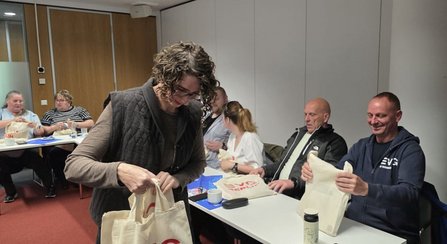  Innenraum mit Tischen und Stühlen. Mehrere Personen sitzen oder stehen an Tischen und halten helle Stofftaschen mit rotem EVG-Logo. Auf den Tischen stehen Becher, Flaschen und Unterlagen. Holzwände und weiße Trennwände im Hintergrund.