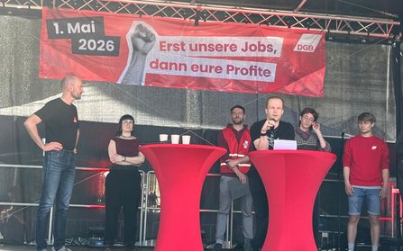 Mehrere Personen stehen auf einer Bühne hinter zwei hohen, roten Stehtischen. Über der Bühne hängt ein rotes Banner mit der Aufschrift „1. Mai 2026“ und „Erst unsere Jobs, dann eure Profite“, daneben ein DGB-Logo und eine Faustgrafik. Auf den Stehtischen stehen Becher und Unterlagen. Die Bühne ist von einer Metallkonstruktion und schwarzem Hintergrund umgeben.