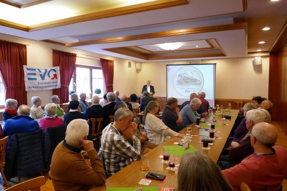 Ein Veranstaltungsraum mit Holztischen und Stühlen, an denen zahlreiche Personen sitzen und nach vorn schauen. Vorn steht eine Person an einem Rednerpult neben einer Projektionsleinwand mit einer runden Abbildung einer Dampflokomotive. An der linken Wand hängt ein Banner mit der Aufschrift „EVG Eisenbahn- und Verkehrsgewerkschaft“. Auf den Tischen stehen Gläser mit Getränken, Untersetzer und kleine Gegenstände. Der Raum hat helle Wände, rote Vorhänge und eine Decke mit Holzverkleidung und eingelassenen Leuchten.