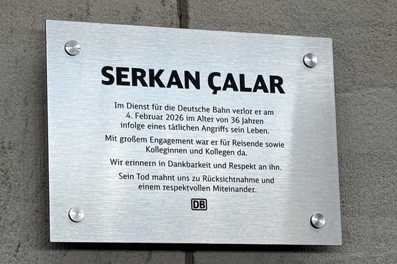 An einer grauen Steinwand ist eine silberne Gedenktafel der Deutschen Bahn angebracht, die an einen Mitarbeiter erinnert, der 2026 bei einem tätlichen Angriff ums Leben kam. Das Bild zeigt eine rechteckige Gedenktafel aus gebürstetem Metall, die mit vier runden Schrauben an einer Steinwand befestigt ist. In großen schwarzen Buchstaben steht oben der Name „SERKAN ÇALAR“. Darunter befindet sich ein Gedenktext der Deutschen Bahn. Der Text erinnert daran, dass die genannte Person am 4. Februar 2026 im Alter von 36 Jahren infolge eines tätlichen Angriffs ums Leben kam. Weiter wird hervorgehoben, dass die Person sich engagiert für Reisende sowie Kolleginnen und Kollegen eingesetzt hat. Die Inschrift schließt mit den Worten, dass der Tod zu Rücksichtnahme und einem respektvollen Miteinander mahne. Am unteren Rand der Tafel ist das Logo der Deutschen Bahn zu sehen. Die Gestaltung ist schlicht und sachlich, mit schwarzer Schrift auf silbernem Hintergrund.