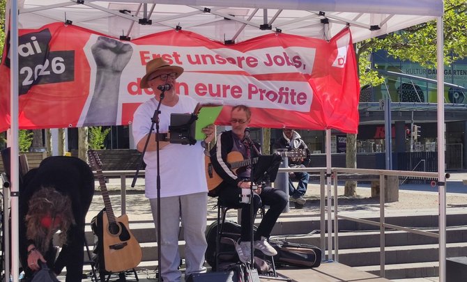 Überdachte Bühne im Freien mit einer Person am Mikrofon, die ein Tablet oder einen Ordner hält, und einer sitzenden Person mit akustischer Gitarre. Links steht eine weitere Gitarre auf einem Ständer. Hinter der Bühne hängt ein rotes Banner mit der Aufschrift „Erst unsere Jobs, dann eure Profite“ und „DGB“. Im Hintergrund sind Bäume, Stufen und ein Gebäude zu sehen.