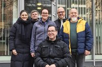 echs Personen vor dem Eingangsbereich eines Gebäudes mit Glasfassade. Fünf Personen stehen, eine Person sitzt in einem Rollstuhl im Vordergrund. Alle tragen Jacken oder Mäntel. Im Hintergrund sind Glastüren, Fenster und Spiegelungen von Gebäuden zu sehen.