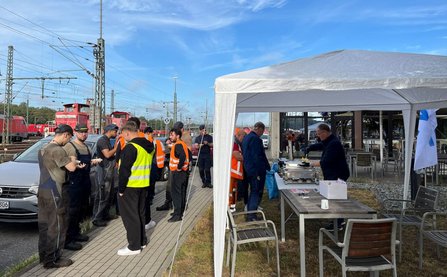 Eine Gruppe von Personen in Arbeitskleidung und Warnwesten steht neben einem weißen Pavillon. Unter dem Pavillon befinden sich Tische mit Servierbehältern und Geschirr. Im Hintergrund sind Bahngleise, Oberleitungen und mehrere rote Lokomotiven unter blauem Himmel zu sehen.