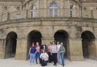 Gruppe von neun Personen steht und kniet vor einem historischen Sandsteingebäude mit mehreren Rundbögen im Erdgeschoss und einer geschwungenen Balustrade im Obergeschoss. Die Personen sind locker nebeneinander angeordnet, eine Person kniet vorne. Hinter der Gruppe befinden sich dunkle Durchgänge unter den Bögen. Im oberen Bereich sind Fenster mit Rundbögen, verzierte Fassadenelemente und geschlossene Sonnenschirme auf der Terrasse sichtbar. Der Boden im Vordergrund ist hell und kiesartig.