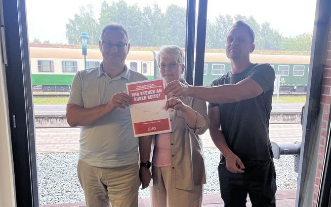 Drei Personen stehen vor einer großen Fensterfront und halten gemeinsam ein rotes und weißes Schild bzw. Dokument in die Kamera. Auf dem Schild steht gut lesbar: „WIR STEHEN AN EURER SEITE!“. Im Hintergrund sind Bahngleise und grün-beige Eisenbahnwagen zu sehen. Der Innenraum wird von Tageslicht durch die Fenster beleuchtet.