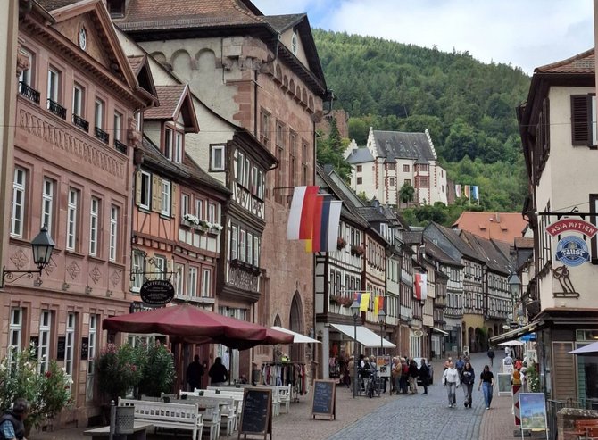 Kopfsteingepflasterte Straße in einer Altstadt mit mehrgeschossigen Fachwerk- und Sandsteingebäuden auf beiden Seiten. Vor den Gebäuden stehen Tische, Bänke und Schilder. Mehrere Fahnen hängen an den Fassaden. Im Hintergrund ist ein bewaldeter Hang mit weiteren Gebäuden sichtbar. Einige Personen gehen auf der Straße.