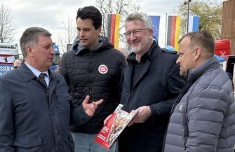Vier Personen stehen im Freien beisammen. Eine Person links spricht zu den drei anderen. Alle tragen Jacken. Eine der Personen hält ein Heft mit rotem Rand und einer bunten Titelseite in den Händen. Im Hintergrund sind mehrere aufgestellte Deutschland- und EU-Flaggen sowie Stände und Fahrzeuge zu sehen. Bäume ohne Laub stehen weiter hinten.
