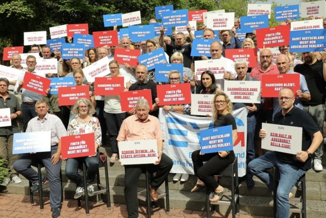 Große Gruppe von Menschen steht im Freien und hält vor sich oder über den Kopf zahlreiche rechteckige Schilder in den Farben Blau, Rot und Weiß. Auf den Schildern sind kurze Slogans in deutscher Sprache zu lesen, jeweils mit „EVG“ als Absender. Einige Personen halten zusätzlich eine blaue Fahne mit weißer Beschriftung. Im Hintergrund sind Pflanzen, ein Gebäude mit heller Fassade und gestapelte Holzscheite zu sehen. Die Personen sind unterschiedlich gekleidet, stehen dicht beieinander und blicken überwiegend zur Kamera.