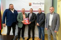 Fünf Personen stehen nebeneinander vor einem weißen Banner. Zwei Personen in der Mitte halten gemeinsam eine rote Broschüre. Auf dem Banner sind das Wappen von Rheinland-Pfalz sowie die Texte „RheinlandPfalz“ und „MINISTERIUM DES INNERN, FÜR INTEGRATION UND VERKEHR“ zu sehen. Die Personen tragen Anzüge, Sakkos oder Hemden. Der Raum hat einen hellen Holzfußboden; links ist ein Teil eines dunklen Tisches sichtbar, rechts eine geöffnete Tür.