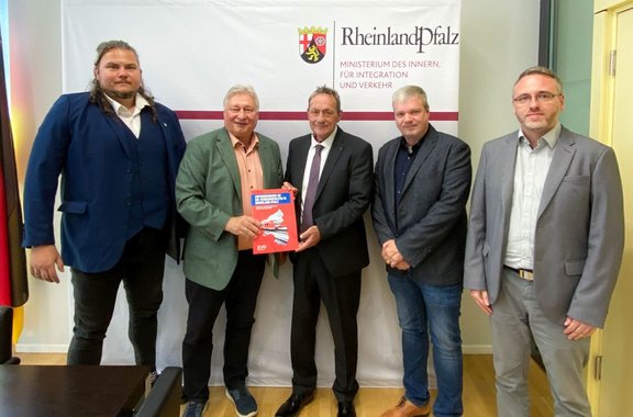 Fünf Personen stehen nebeneinander vor einem weißen Banner. Zwei Personen in der Mitte halten gemeinsam eine rote Broschüre. Auf dem Banner sind das Wappen von Rheinland-Pfalz sowie die Texte „RheinlandPfalz“ und „MINISTERIUM DES INNERN, FÜR INTEGRATION UND VERKEHR“ zu sehen. Die Personen tragen Anzüge, Sakkos oder Hemden. Der Raum hat einen hellen Holzfußboden; links ist ein Teil eines dunklen Tisches sichtbar, rechts eine geöffnete Tür.