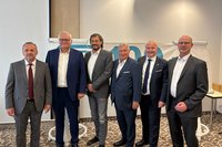 Sechs Personen in Business-Kleidung stehen nebeneinander vor einem Banner in einem Konferenz- oder Tagungsraum und blicken in die Kamera. Die sechs Personen sind in einer Reihe aufgestellt und tragen überwiegend Anzüge mit Hemd und teilweise Krawatte. Hinter ihnen hängt ein weißes Banner mit großen blau dargestellten Ziffern, von denen nur ein Teil sichtbar ist. Links und rechts des Banners stehen Flipcharts. Der Raum hat helle Wände, eine hohe Decke und einen gemusterten Teppichboden. Die Aufnahme zeigt die Personen von etwa den Knien aufwärts.