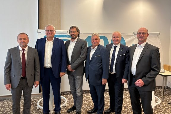 Sechs Personen in Business-Kleidung stehen nebeneinander vor einem Banner in einem Konferenz- oder Tagungsraum und blicken in die Kamera. Die sechs Personen sind in einer Reihe aufgestellt und tragen überwiegend Anzüge mit Hemd und teilweise Krawatte. Hinter ihnen hängt ein weißes Banner mit großen blau dargestellten Ziffern, von denen nur ein Teil sichtbar ist. Links und rechts des Banners stehen Flipcharts. Der Raum hat helle Wände, eine hohe Decke und einen gemusterten Teppichboden. Die Aufnahme zeigt die Personen von etwa den Knien aufwärts.