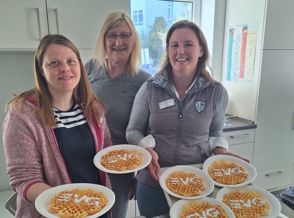 Drei Personen stehen in einer hellen Küche und halten jeweils weiße Pappteller mit herzförmigen Waffeln. Auf den Waffeln sind mit weißem Zuckerguss Buchstaben aufgebracht. Insgesamt sind sechs Waffeln sichtbar, einige werden einzeln, andere zu zweit auf einem Teller gehalten. Im Hintergrund sind weiße Küchenschränke, eine Arbeitsfläche, ein Fenster mit Blick nach draußen und ein farbiger Aushang an der Wand zu sehen.