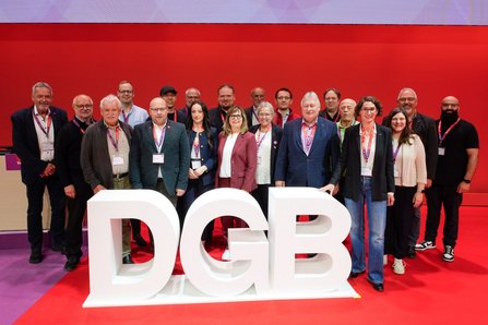 Gruppenfoto mehrerer Personengruppe in ähnlicher Anordnung. Die großen weißen Buchstaben „DGB“ stehen im Vordergrund auf dem Boden. Die Personen stehen dicht nebeneinander vor einer roten Bühnenwand.