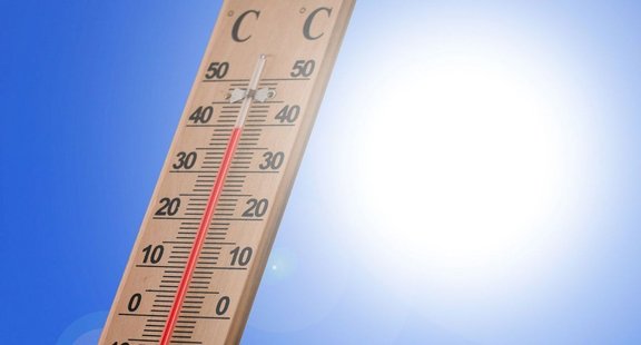 Holzthermometer vor blauem Himmel, die rote Flüssigkeit steht zwischen etwa 35 und 40 Grad Celsius, Sonnenlicht mit Lensflare sichtbar.