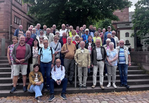 Große Gruppe von etwa 40 Personen auf einer breiten Steintreppe vor einem mehrstöckigen Gebäude aus rotem Sandstein. Hinter der Gruppe steht ein großer Baum mit dichtem Laub. Die Personen sind in mehreren Reihen angeordnet und tragen überwiegend Freizeitkleidung in verschiedenen Farben. Im Vordergrund befinden sich Pflastersteine und trockenes Laub.