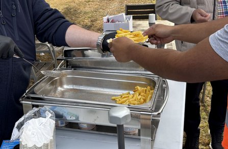 Nahaufnahme eines Speisenausgabebehälters aus Edelstahl. Eine Person mit Handschuh hält eine Schöpfkelle über dem Behälter, während eine andere Person einen Pappteller mit Pommes frites entgegennimmt. Im Hintergrund stehen ein Tisch, eine Box mit Portionsbeuteln und weitere Personen.