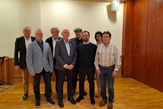 Gruppenfoto von acht Personen, die nebeneinander in einem Raum mit Holzboden stehen. Die Personen tragen unterschiedliche Kleidung wie Jacken, Hemden und Pullover. Im Hintergrund ist eine helle Wand mit einem Wandlautsprecher sowie eine Holztür sichtbar. Die Personen stehen dicht beieinander und blicken in Richtung der Kamera.