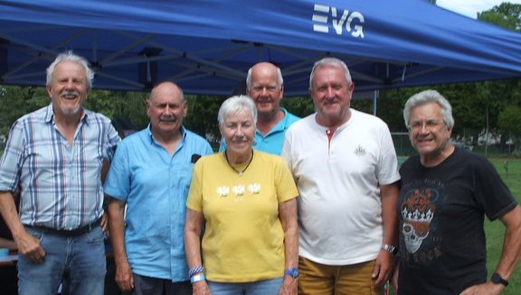 Sechs Personen stehen dicht nebeneinander vor einem blauen Pavillon mit dem weißen Schriftzug „EVG“. Die Personen tragen unterschiedlich farbige Freizeitkleidung, darunter karierte, blaue, gelbe, weiße und dunkle Oberteile. Im Hintergrund sind Bäume und eine Grünfläche zu sehen.