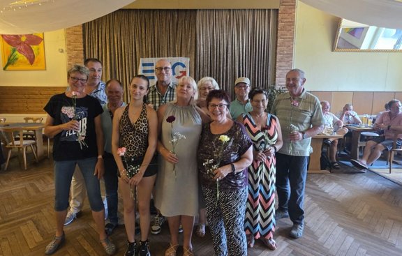 Gruppe von elf Personen steht in einem Saal vor einem Vorhang und einem Banner mit dem Schriftzug „EVG“. Mehrere Personen halten einzelne Blumen in den Händen. Im Hintergrund stehen Tische und Stühle, an denen weitere Personen sitzen. Die Decke ist mit hellen Stoffbahnen dekoriert.