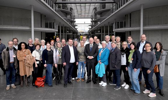 Große Gruppe von etwa 30 bis 40 Personen steht dicht nebeneinander in einem modernen, mehrstöckigen Innenhof mit grauen Betonwänden, umlaufenden Galerien und Glasdach. Die Personen sind frontal zur Kamera ausgerichtet; Gesichter sind unscharf dargestellt. Die Gruppe trägt überwiegend Alltags- und Businesskleidung in dunklen und neutralen Farben, einzelne Kleidungsstücke in Blau, Grün oder Braun fallen auf. Im Vordergrund ist ein dunkler Steinboden zu sehen, im Hintergrund die symmetrische Architektur mit Geländern, Säulen und oberen Ebenen. Licht fällt gleichmäßig von oben durch das Glasdach ein.