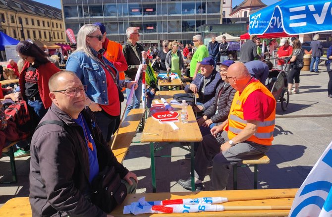 Blick über mehrere Biertischgarnituren mit sitzenden und stehenden Personen. Mehrere Personen tragen orangefarbene „EVG“-Warnwesten. Auf einem Tisch liegen Schilder mit „EVG“. Im Hintergrund sind ein großes Gebäude mit Glasfassade, weitere Stände, Fahnen und Menschen auf dem Platz zu sehen.