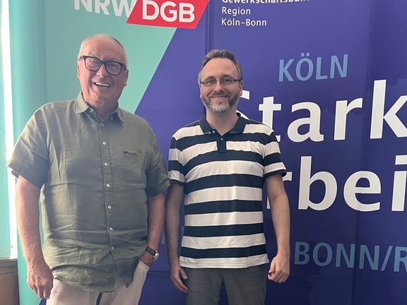 Zwei Personen stehen nebeneinander vor einer großen, bunten Wand mit der Aufschrift „DGB Region Köln-Bonn“ und „Köln“. Die Aufnahme zeigt zwei Erwachsene von etwa gleicher Körpergröße, die nebeneinander stehen und in die Kamera schauen. Die Person links trägt ein kurzärmeliges, einfarbiges Hemd und eine helle Hose sowie eine Brille. Die Person rechts trägt ein gestreiftes Oberteil mit kurzen Ärmeln und ebenfalls eine Brille. Beide stehen aufrecht und nah beieinander. Im Hintergrund befindet sich eine großformatige Wand oder ein Banner mit mehreren Farben, darunter Grün, Blau und Rot. Darauf sind gut lesbare Texte zu sehen, unter anderem „DGB NRW“, „Region Köln-Bonn“ sowie „Köln“. Ein Fenster am linken Bildrand lässt Tageslicht in den Raum, wodurch die Szene gleichmäßig beleuchtet ist.