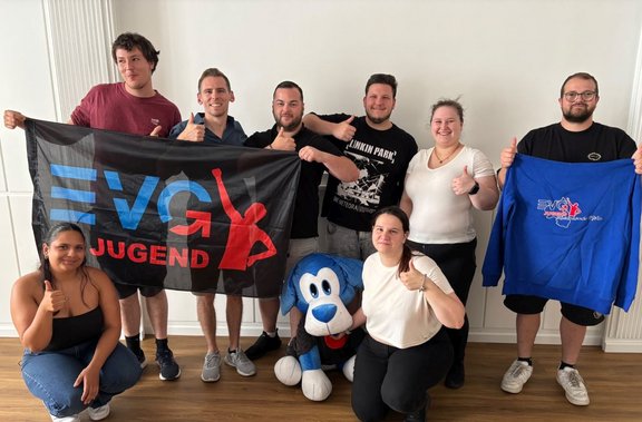 Gruppe von acht Personen in einem Innenraum mit weißen Wänden und Holzfußboden. Vier Personen stehen in der hinteren Reihe, zwei sitzen vorn auf dem Boden, eine Person kniet rechts vorn und eine steht rechts außen. Zwei Personen halten eine schwarze Fahne mit der Aufschrift „EVG Jugend“ und roter Silhouette. Rechts hält eine Person ein blaues Sweatshirt mit einem ähnlichen Logo. Mehrere Personen zeigen einen Daumen nach oben. In der Mitte am Boden liegt ein großes blaues Plüschtier in Form eines Hundes.