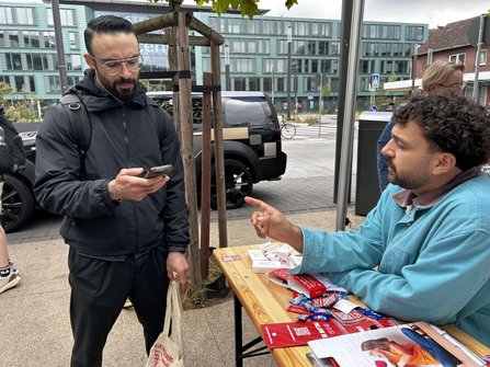 Außenbereich mit einem Holztisch unter einem Pavillon. Auf dem Tisch liegen rote Flyer, Karten, Süßigkeiten und ein Klemmbrett mit Unterlagen. Eine Person steht vor dem Tisch und hält ein Smartphone sowie eine helle Stofftasche. Gegenüber sitzt eine Person an dem Tisch und zeigt mit einem Finger auf das Smartphone. Im Hintergrund sind ein geparktes Fahrzeug, junge Bäume und mehrgeschossige Gebäude zu sehen.