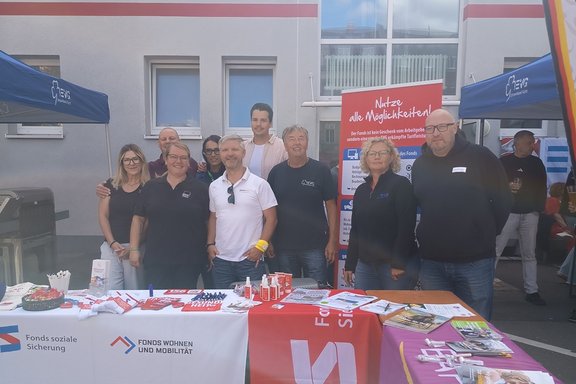 Mehrere Personen stehen an einem Informationsstand im Freien hinter Tischen mit Broschüren und Werbematerialien. Das Foto zeigt eine Gruppe von neun Personen, die vor einem Gebäude an einem Informationsstand für soziale Leistungen und Mobilitätsangebote posieren. Die Personen stehen hinter mehreren aneinander gereihten Tischen, die mit Informationsmaterial, Flyern, Kugelschreibern, Werbeartikeln und weiteren Auslagen bedeckt sind.