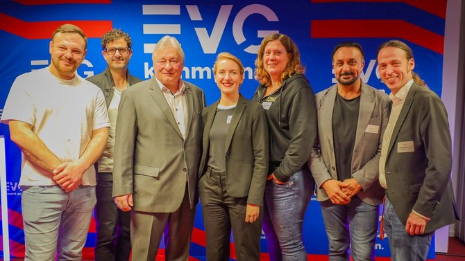 Eine Gruppe von sieben Personen steht nebeneinander vor einer Wand mit blauem Hintergrund und weiß-roten grafischen Elementen sowie dem gut sichtbaren Schriftzug „EVG“. Die Personen blicken zur Kamera. Einige tragen helle oder dunkle T‑Shirts, andere Jacketts oder Hoodies. Die Personen stehen dicht beieinander, teilweise mit gefalteten Händen oder Händen in den Taschen.