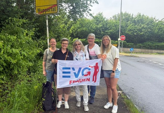 Fünf Personen stehen nebeneinander am Straßenrand vor einem Ortsschild mit der Aufschrift „Wuppertal“ und halten ein weißes Banner mit dem blauen Schriftzug „EVG“ und rotem Zusatz „FRAUEN“. Das Banner zeigt zusätzlich eine rot dargestellte Figur mit ausgestrecktem Arm. Die Personen tragen Freizeitkleidung wie T-Shirts, kurze Hosen, Jeans und Sneakers; einige tragen Brillen und haben Schultertaschen oder Rucksäcke dabei. Im Hintergrund sind eine asphaltierte Straße, ein Stoppschild, ein Verkehrszeichen mit Pfeil, eine Leitplanke sowie dichter grüner Bewuchs mit Bäumen und Sträuchern zu sehen. Die Szene spielt im Freien bei Tageslicht.