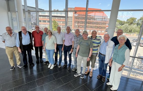 Gruppe von 13 Personen steht nebeneinander in einem hellen Innenraum mit großen Fensterfronten. Die Personen tragen überwiegend Freizeitkleidung in verschiedenen Farben, darunter Hemden, Poloshirts, T-Shirts, Jeans und helle Hosen. Durch die Fenster sind mehrere Gebäude, Bäume, parkende Fahrzeuge und ein sonniger Himmel sichtbar. Der Boden besteht aus dunklen, glänzenden Fliesen.