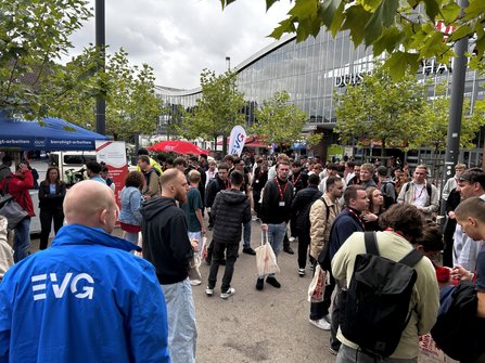 Außenbereich vor einem großen Gebäude mit gebogener Glasfassade und teilweise sichtbarer Beschriftung „DUIS…“. Zwischen mehreren Bäumen stehen zahlreiche Personen auf einem Platz. Im Vordergrund ist eine Person mit blauer Jacke und weißem „EVG“-Aufdruck auf dem Rücken zu sehen. Links stehen ein blaues Zelt und mehrere Fahnen sowie Werbebanner. Viele Personen tragen Umhängetaschen oder Namensschilder. Der Himmel ist bewölkt.