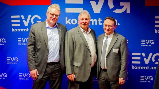 Drei Personen stehen nebeneinander vor einer blauen Wand mit mehrfach sichtbarem EVG‑Logo und dem Schriftzug „kommt an.“. Alle tragen formelle Kleidung und Namensschilder. Die Person links trägt eine Brille und einen Anzug mit offenem Hemdkragen. Die Person in der Mitte trägt einen Anzug mit hellem Hemd. Die Person rechts trägt einen Anzug mit Krawatte.
