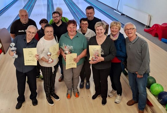 Gruppenfoto in einer Bowlinghalle: Etwa 14 Personen stehen dicht nebeneinander auf der Holzbahn vor mehreren Bowlingbahnen. Die Gesichter sind unscharf dargestellt. Mehrere Personen halten Urkunden aus gelbem Papier, kleine Pokale oder verpackte Präsente in den Händen. Im Hintergrund sind die Bowlingbahnen mit Pins zu sehen, rechts am Rand liegen mehrere Bowlingkugeln auf einer Ablage. Die Personen tragen Freizeitkleidung in unterschiedlichen Farben, einige stehen in einer vorderen, andere in einer hinteren Reihe.