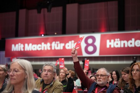 Blick in einen Saal mit sitzendem Publikum. Mehrere Personen halten rote Karten mit weißem Aufdruck nach oben. Im Hintergrund hängt ein langes rotes Banner mit weißem Text, teilweise lesbar „Mit Macht für die … Hände“ sowie eine große weiße Zahl „8“.