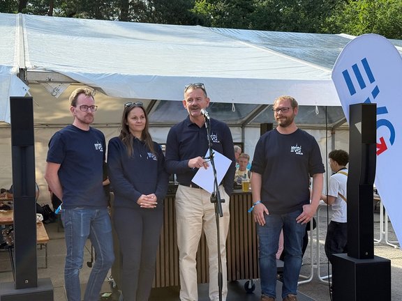 Vier Personen stehen gemeinsam auf einer kleinen Bühne vor einem Zelt; eine Person spricht an einem Mikrofon und hält ein Blatt Papier. Die vier Personen stehen nebeneinander im Vordergrund. In der Mitte steht eine Person an einem Mikrofonständer und hält ein weißes Blatt in der Hand. Alle vier Personen tragen dunkle T-Shirts oder Oberteile mit einem kleinen weißen Logo auf der Brust. Links und rechts neben der Gruppe stehen schwarze Lautsprecher. Hinter der Gruppe ist ein großes, helles Zelt aufgebaut, darunter sind Tische und weitere Personen zu sehen. Rechts im Bild steht eine hohe, schmale Fahne mit blauem und rotem Aufdruck. Im Hintergrund sind grüne Bäume und Tageslicht sichtbar.
